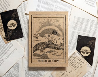 May include: A collection of vintage-style tarot cards. The central card features an illustration of a woman with a cup, the words "QUEEN OF CUPS" are printed below. Other cards show a moth and celestial designs. The cards are on a background of aged paper.