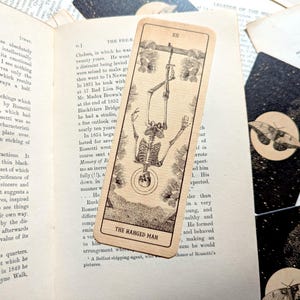 Puede incluir: Una carta de tarot vintage con una ilustración de un esqueleto colgado boca abajo, titulada "THE HANGED MAN". La carta está colocada sobre un libro abierto, con otras cartas con diseños de luna y polilla.