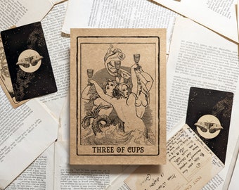 May include: A tarot card with the text "THREE OF CUPS" features an illustration of three figures raising chalices. The card is surrounded by other cards and vintage paper ephemera, creating a mystical aesthetic.