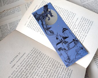 May include: A rectangular bookmark with a periwinkle blue background featuring a black ink illustration of a dragon flying toward a full moon above a castle. The design includes small gold-colored star accents scattered across the night sky.