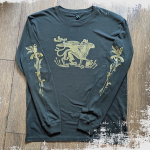 May include: Dark olive green long-sleeve shirt with a gold griffin graphic on the front and gold floral sword designs on the sleeves. The shirt has a crew neck and is made of a soft material.