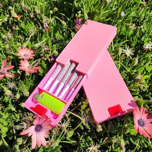 May include: A pink box with a hinged lid, open to reveal several pre-rolled cigarettes and a green lighter. The box is resting on a bed of green grass and colorful flowers, with the lid partially open.