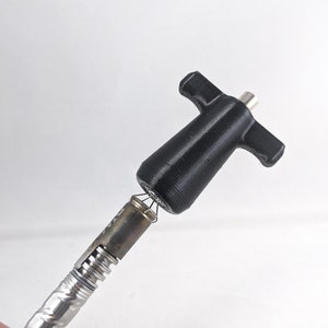 Puede incluir: Una herramienta de plástico negro con un mango en forma de T y una punta de metal roscada. La herramienta está unida a una bobina de metal plateado.