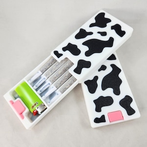 Può includere: Una custodia a stampa mucca bianca e nera con una chiusura rosa. La custodia è aperta e contiene quattro sigarette pre-rollate e un accendino verde. Le sigarette sono etichettate "Elements".