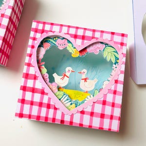 May include: A collection of handmade paper art boxes. One box features a heart-shaped cutout with two ducks, another with two cats, and a third with two dogs. The boxes have a pink and white gingham pattern.