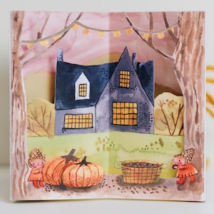 Peut inclure: Illustration à l'aquarelle d'une maison au toit bleu foncé, entourée d'arbres et d'un ciel rose. Deux citrouilles et un panier sont devant la maison. Deux petits personnages en tenues orange se tiennent de chaque côté.