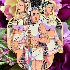 May include: A gold-toned enamel pin featuring three figures in white and pink outfits with gold accents. The central figure holds a sword with a pink and blue blade. The characters have intricate hair and skin patterns, set against a dark, arched background with gold lines.