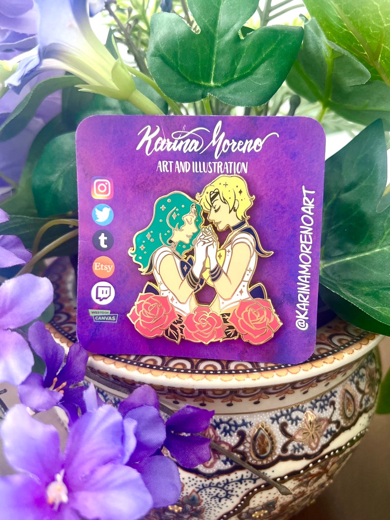 Puede incluir: Pin de esmalte dorado con dos personajes de anime, una mujer con cabello verde y un hombre con cabello rubio, tomados de la mano y de pie frente a tres rosas rojas. El pin tiene un fondo morado con el texto "Karina Moreno Art and Illustration" y "@karinamorenoart".