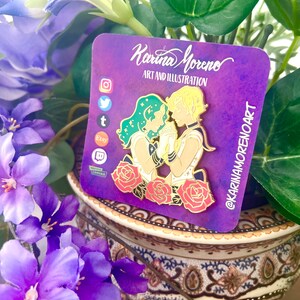 Puede incluir: Un pin de esmalte dorado con dos figuras, una con cabello verde y la otra con cabello amarillo, tom&aacute;ndose de la mano. Las figuras est&aacute;n rodeadas de rosas rojas. El pin est&aacute; sobre un fondo morado con el texto "Karina Moreno Art and Illustration" y "@karinamorenoart".