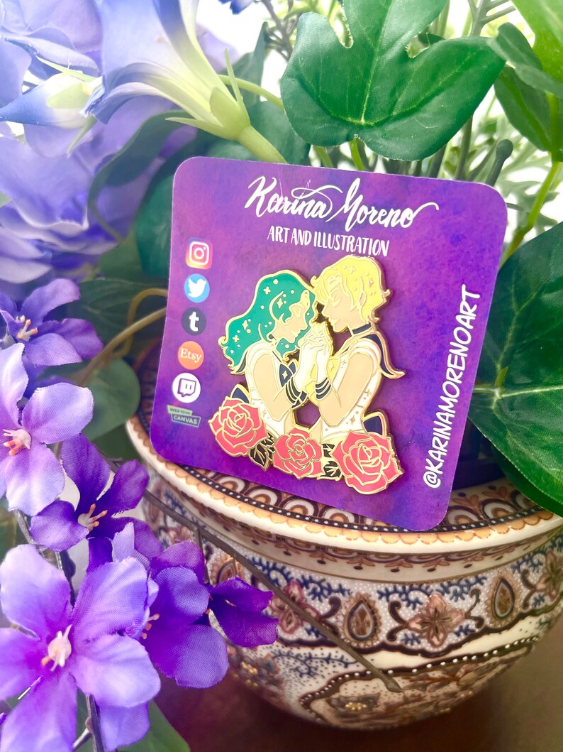 Puede incluir: Un pin de esmalte dorado con dos figuras, una con cabello verde y la otra con cabello amarillo, tom&aacute;ndose de la mano. Las figuras est&aacute;n rodeadas de rosas rojas. El pin est&aacute; sobre un fondo morado con el texto "Karina Moreno Art and Illustration" y "@karinamorenoart".