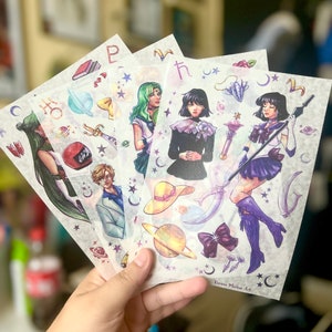 May include: Four clear sticker sheets with colorful illustrations of anime characters and celestial objects. The characters are wearing outfits with a variety of colors, including blue, green, purple, and black. The stickers are perfect for adding a touch of whimsy to laptops, water bottles, or journals.