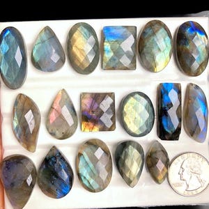 Pode incluir: Uma coleção de pedras preciosas de labradorita facetadas em várias formas, incluindo ovais, em forma de gota e retangulares. As pedras exibem flashes iridescentes de azul, verde e dourado. Uma moeda é incluída para referência de tamanho.