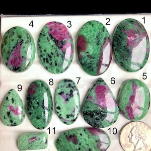 Könnte beinhalten: Eine Sammlung polierter Rubin-Zoisit-Edelsteine in verschiedenen Formen, darunter oval, tropfenförmig und rechteckig. Die Steine zeigen eine Mischung aus grünen und rosa Farbtönen mit schwarzen Sprenkeln. Eine Münze dient als Größenreferenz.