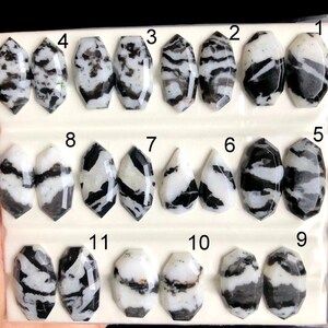May include: A collection of eleven polished zebra jasper gemstones, each with a unique pattern of black and white swirls and stripes. The stones vary in shape, including oval, teardrop, and faceted designs. The gemstones are displayed on a white surface.