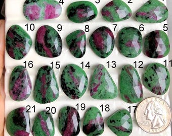 May include: A collection of twenty-one ruby zoisite gemstones, each with a unique shape and faceting. The stones display a mix of green, black, and pink hues. A US quarter is included for size reference.