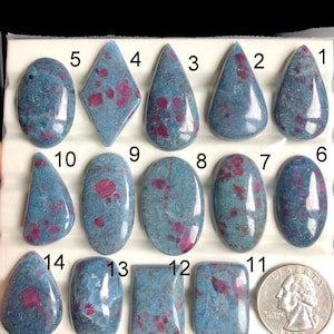 May include: A collection of fourteen polished gemstones in various shapes, including oval, teardrop, and rectangular. The stones feature a blue-green matrix with red inclusions. A US quarter is included for size reference.