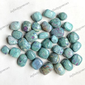 May include: A collection of tumbled stones in shades of teal, green, and blue, with flecks of red. The stones vary in size and shape, and are scattered across a white surface. The overall aesthetic is natural and organic.