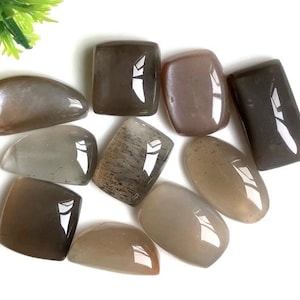 Può includere: Una collezione di dieci cabochon di pietra di sole in varie tonalità di grigio e marrone. Le pietre sono lisce e levigate, con un leggero luccichio.