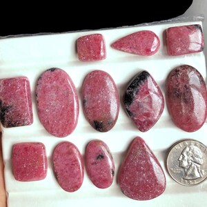 May include: Assortment of polished rhodochrosite gemstones in various shapes, including rectangles, ovals, and a teardrop. The stones display a vibrant pink hue with darker veining and markings. A US quarter coin is included for size reference.