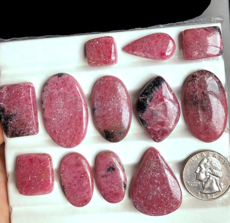 May include: Assortment of polished rhodochrosite gemstones in various shapes, including rectangles, ovals, and a teardrop. The stones display a vibrant pink hue with darker veining and markings. A US quarter coin is included for size reference.