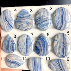 May include: A collection of eleven polished gemstones with a blue and white banded pattern. The stones vary in shape, including oval, teardrop, and rectangular. The gemstones are displayed on a white background.