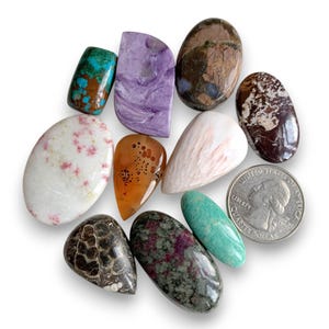 May include: A collection of polished gemstones in various shapes and colors, including turquoise, purple, brown, white, orange, and green. A U.S. quarter coin is included for size reference. The gemstones are arranged on a white surface.