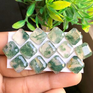 Op de afbeelding: Een verzameling van 12 groene mosagaat-cabochons, elk met een uniek patroon van groen en wit. De cabochons zijn vierkant gesneden en hebben een gladde, gepolijste afwerking.