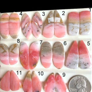 Puede incluir: Colección de piedras preciosas de ópalo rosa en varias formas, incluyendo ovaladas, de lágrima y rectangulares. Los ópalos muestran una gama de tonos rosados, algunos con vetas blancas y grises. Se incluye una moneda para referencia de tamaño.