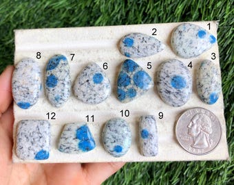 May include: Twelve blue and white cabochon stones with a speckled pattern. The stones are arranged in rows on a white card with numbers 1 through 12 printed on the card. A US quarter is included for scale.