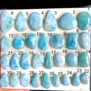 May include: A collection of thirty polished larimar gemstones, each with unique patterns and shades of blue and white. The gemstones are displayed on a white surface, numbered for identification. The shapes vary from oval to teardrop.