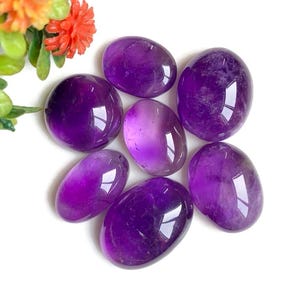 Peut inclure: Sept pierres d'améthyste polies de différentes nuances de violet, du violet foncé au lavande clair. Les pierres lisses de forme ovale reflètent la lumière, créant un aspect brillant. Quelques feuilles vertes et fleurs oranges sont dans le coin supérieur gauche.