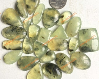 May include: A collection of light green prehnite gemstones in various shapes, including ovals, teardrops, and rectangles. The stones have black inclusions and are displayed with a quarter for size reference.