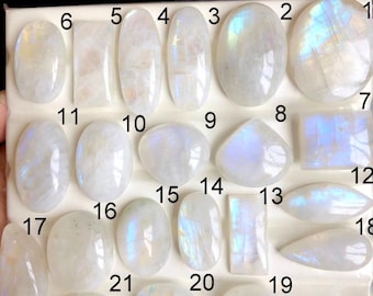 May include: A collection of polished moonstone gemstones in various shapes and sizes, including oval, rectangular, and teardrop. The stones display a milky white base with iridescent blue and rainbow flashes. The gemstones are arranged on a white surface.