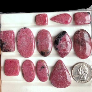 May include: A collection of polished rhodochrosite gemstones in various shapes, including rectangles, ovals, and teardrops. The stones display a vibrant pink hue with darker veining and markings. A U.S. quarter is included for size reference.