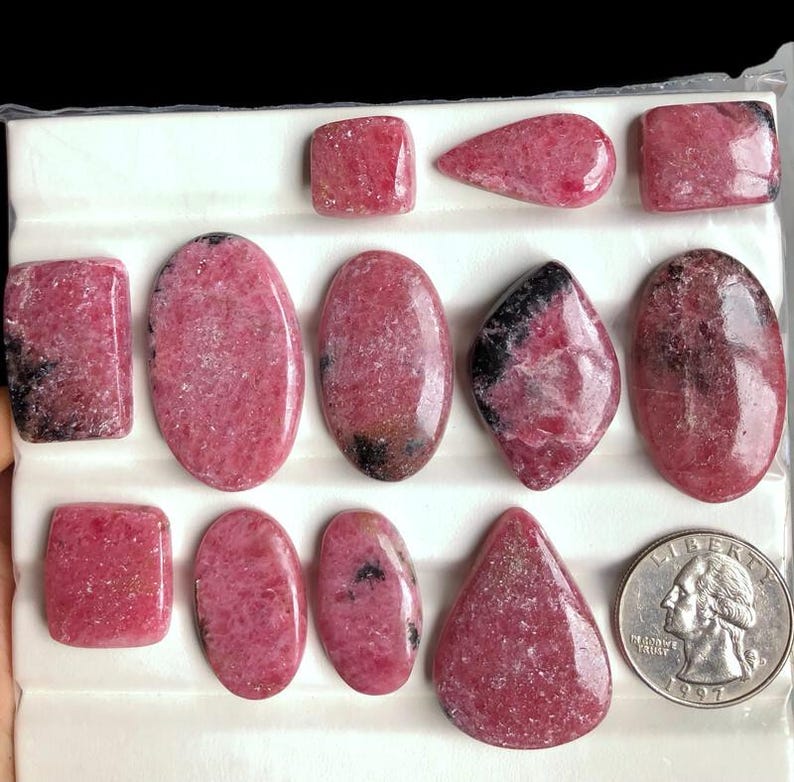 May include: A collection of polished rhodochrosite gemstones in various shapes, including rectangles, ovals, and teardrops. The stones display a vibrant pink hue with darker veining and markings. A U.S. quarter is included for size reference.