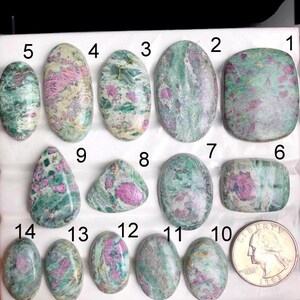 May include: A collection of fourteen polished gemstones, each with a unique shape and pattern. The stones display a mix of green and pink hues, with some featuring a speckled or marbled appearance. A US quarter is included for size reference.