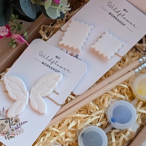 May include: A craft kit featuring white butterfly and square-shaped earrings, paint pots, and paintbrushes. The kit includes a card with the text "MY Wildflower WORKSHOP". The kit is presented in a brown box with decorative paper and flowers.