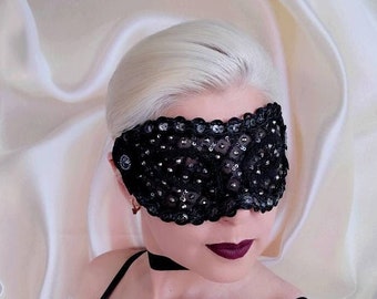 May include: A black sleep mask with a delicate lace design and sparkling sequins. The mask is worn by a person with blonde hair and dark lipstick.