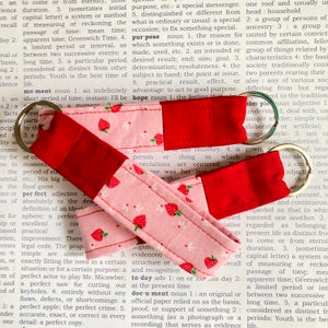 May include: Two fabric keychains with a strawberry pattern. One keychain is red and the other is pink with white polka dots. The keychains are made of fabric and have a metal ring.