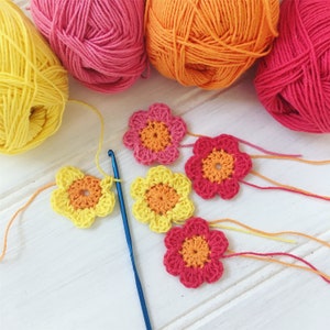 Puede incluir: Cinco motivos de flores de ganchillo en tonos de rosa, amarillo y naranja. Las flores est&aacute;n dispuestas sobre una superficie blanca con un gancho de ganchillo azul.