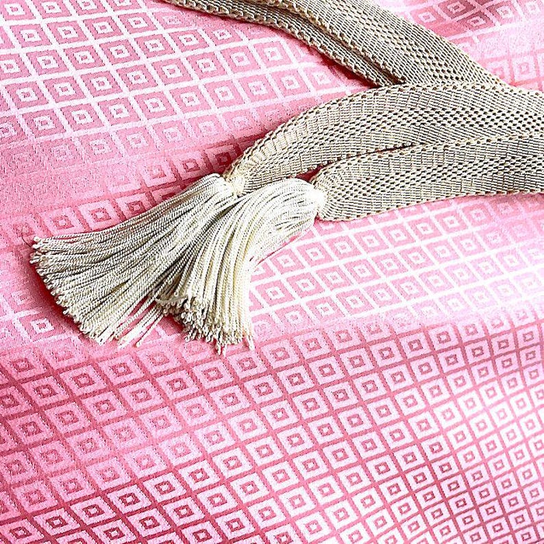 May include: A pink fabric with a white geometric pattern and a white tassel. The fabric has a gradient effect, with the pink color fading to white.