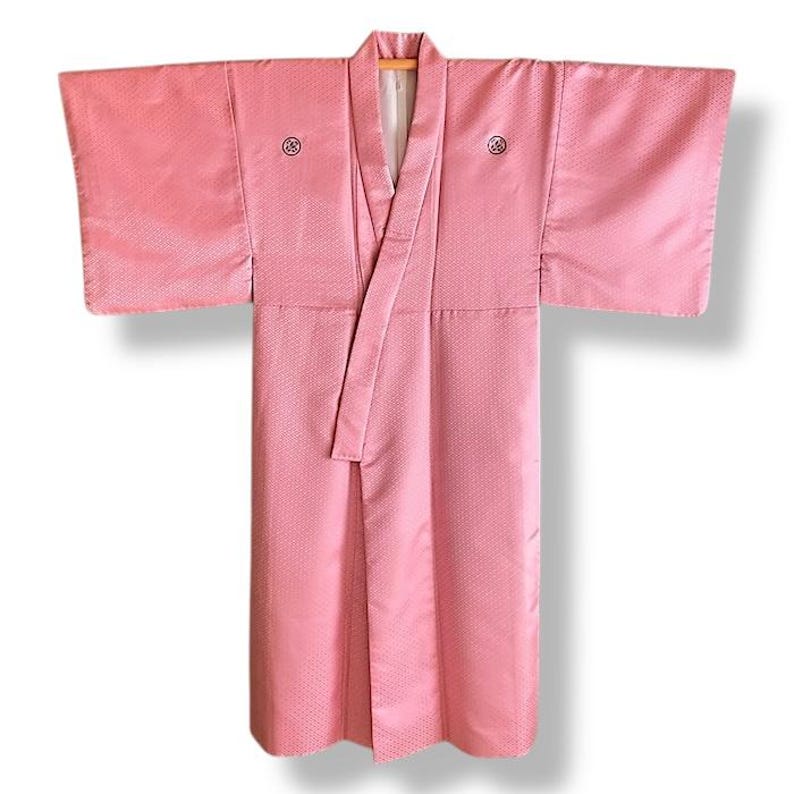 May include: A pink Japanese kimono with a white collar and a small black design on the front. The kimono is made of a silky fabric and has a traditional style.