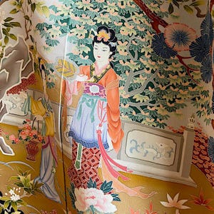 Puede incluir: Un kimono japonés tradicional con un estampado floral colorido que presenta a una mujer en un entorno de jardín. El kimono está hecho de seda y tiene un fondo dorado con detalles intrincados.