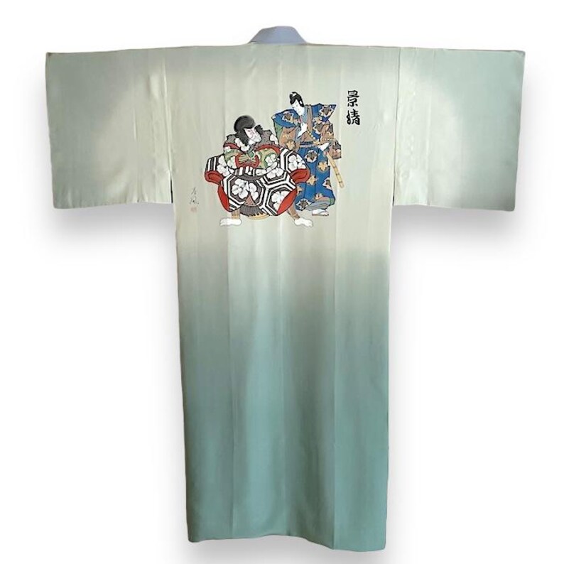 Puede incluir: Un kimono japon&eacute;s de color verde claro y blanco degradado con una escena tradicional japonesa de dos figuras con ropa colorida impresa en la parte posterior. El texto "歌舞伎" est&aacute; impreso en el lado derecho de la imagen.
