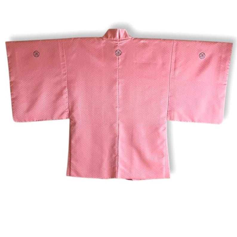 May include: A pink Japanese haori, a traditional short jacket, with a subtle pattern and three black embroidered circles on the back.