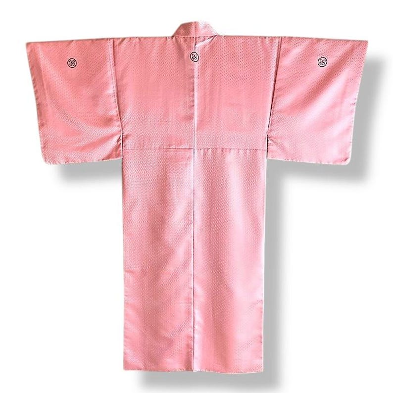 May include: A pink Japanese kimono with a traditional pattern. The kimono has a high collar and long sleeves. There are three small black circles on the back of the kimono.