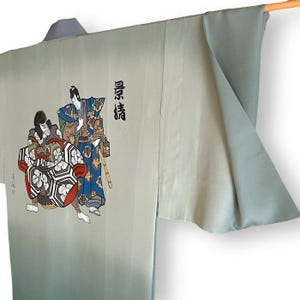 Puede incluir: Un kimono azul claro con un efecto degradado descolorido. El kimono presenta un dise&ntilde;o pintado a mano de dos figuras con ropa japonesa tradicional. El texto "真情" est&aacute; escrito en negro en el lado derecho del kimono.