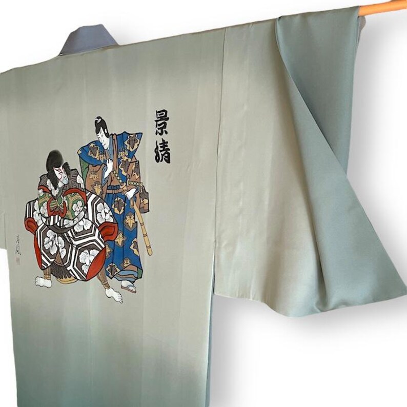 Puede incluir: Un kimono azul claro con un efecto degradado descolorido. El kimono presenta un dise&ntilde;o pintado a mano de dos figuras con ropa japonesa tradicional. El texto "真情" est&aacute; escrito en negro en el lado derecho del kimono.