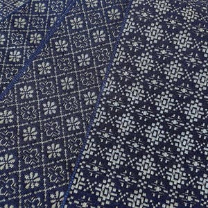 May include: Close-up of several navy blue placemats with a white geometric pattern. The placemats have a woven texture and a repeating design of floral and square shapes. The edges are trimmed with a darker blue border.