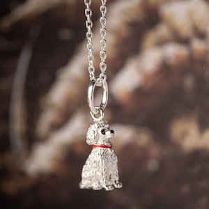Puede incluir: Un colgante de metal plateado con forma de perro caniche sentado, que presenta un pequeño collar rojo alrededor del cuello. El colgante está suspendido de una cadena de eslabones de metal plateado a juego, mostrando una textura de pelaje detallada.